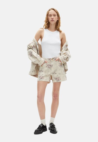 Camiseta sin mangas blanca de rayas, pantalones cortos beiges con estampado floral y una chaqueta a juego. La modelo lleva puestos zapatos negros y calcetines blancos.