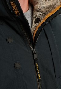 Zwarte jas met een zachte, structuurgebonden kraag en gele accenten. Voorzien van een ritssluiting aan de voorkant en knoopdetails. Merkenlogo zichtbaar.