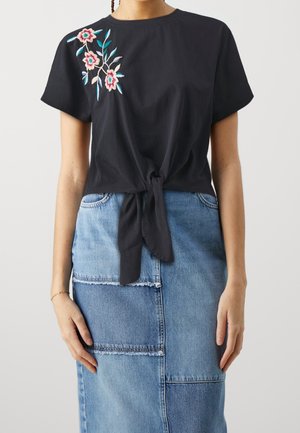Top nero a maniche corte con ricamo floreale nei toni del rosa e dell'azzurro, con un nodo in vita. Indossato con una gonna di jeans.