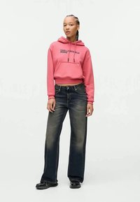 Mulher vestindo um hoodie rosa com a inscrição "Karl Lagerfeld Jeans", jeans de perna larga pretos desbotados e sapatos pretos, em pé contra um fundo simples.