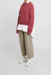 Pull en tricot d'un rose poussiéreux, avec une texture côtelée. Porté par-dessus une chemise blanche à col, associé à un pantalon large beige et des baskets blanches.