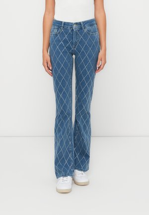 Blauwe flare jeans met een diamanten patroon in de steek, vijf zakken en een sluiting met een knop aan de voorkant. Gecombineerd met witte sneakers.