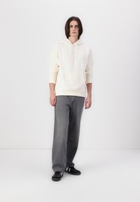 Hanorac crem cu glugă, cu buzunar frontal, asortat cu blugi gri cu picior larg și adidași negri cu accente albe. Design minimalist.
