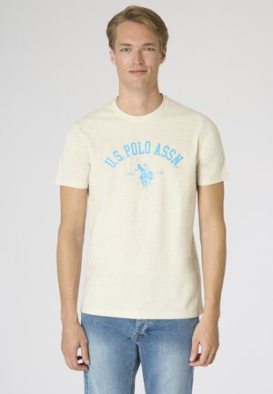 Giovane con capelli chiari che indossa una maglietta color crema U.S. Polo Assn. e jeans blu, in piedi davanti a uno sfondo chiaro e semplice.