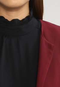 Black high-neck top with ruffled neckline, paired with a maroon blazer. The fabrics appear smooth with a tailored design and defined edges.
