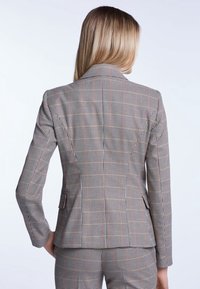Blazer gris à carreaux avec une coupe ajustée, deux poches avant et de subtils accents rouges. Tissu lisse et épaules structurées, vu de dos.