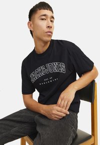 Sort bomulds t-shirt med hvid "JACK & JONES" logo. Korte ærmer, rund hals, afslappet pasform. Modellens siddende på en træstol.