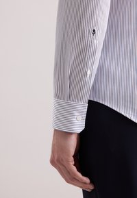 Close-up van de arm van een persoon die een wit met blauw gestreept overhemd met lange mouwen draagt, met knopen aan de manchetten en een kleine zwart geborduurde roos op de mouw.