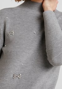 Maglione grigio a coste con accenti di gioielli e fiocchi decorativi, con una finitura testurizzata. Collo alto e maniche aderenti.