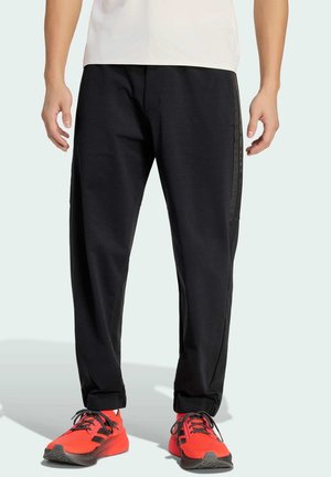 Mężczyzna w czarnych casualowych spodniach i jasnoczerwonych sportowych butach, stojący na tle jednolitego jasnego tła.