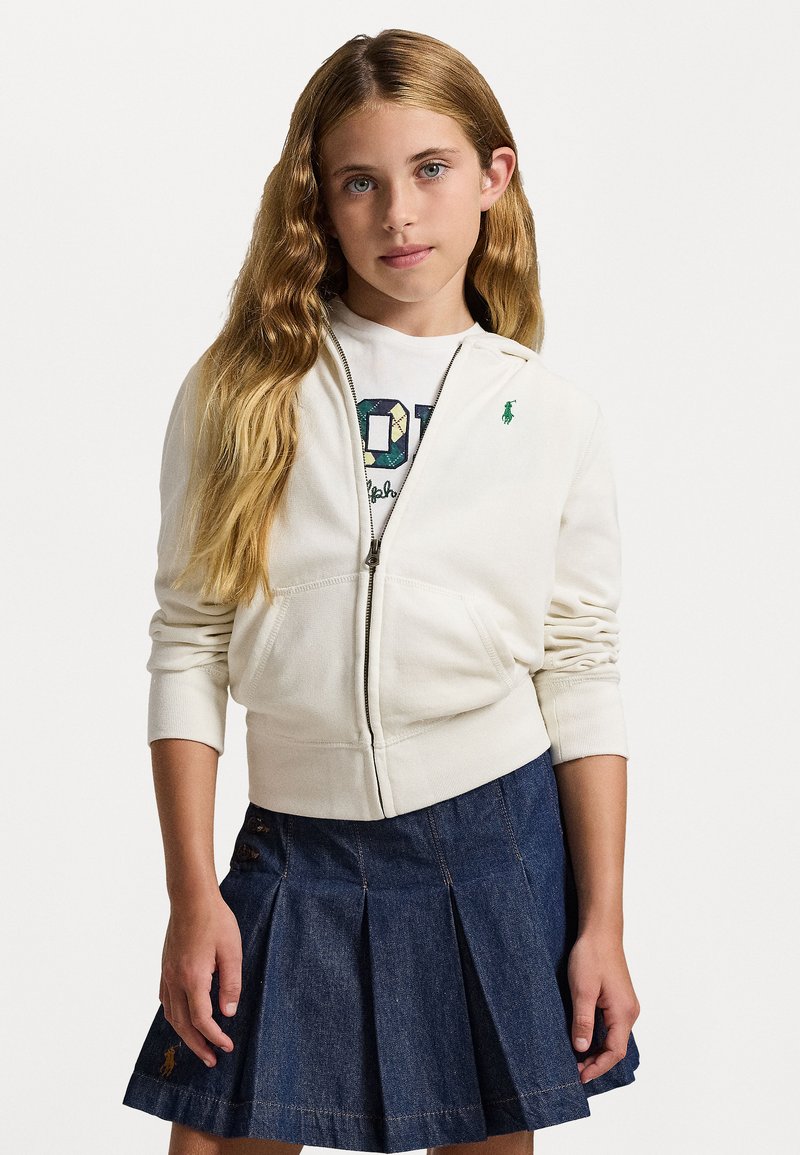 Bijela zip-up hoodie sa prednjim džepovima i zelenim logom, uparen s plavom traper suknjom s plisiranim detaljima i dekorativnim gumbima.