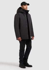 Giacca nera con cappuccio e texture liscia, chiusura frontale con zip e un patch con logo all'orlo, abbinata a jeans scuri e stivali.