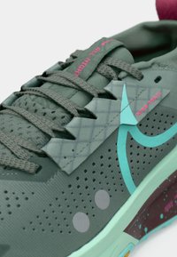 Közelkép egy zöld Nike Trail futócipőről, türkiz szwoosh-sal, perforált felsőrésszel, rózsaszín "ALL NIGHT" és "NIKE TRAIL" felirattal, valamint pettyezett középtalppal.