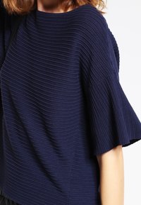 Nainen, jolla on yllään tummansininen ribbiknitti, jossa on lyhyet liehutetut hihat, näkyy niskasta vyötärölle valkoista taustaa vasten.