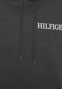 Sudadera gris oscuro hecha de tela suave. Presenta un logo blanco de "HILFIGER" en el pecho y cordones negros con puntas metálicas.