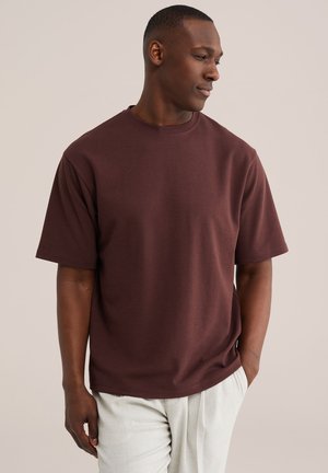 Uomo che indossa una t-shirt a maniche corte marrone scuro e pantaloni chiari, che guarda verso destra con una mano in tasca.