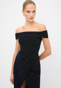 Abito nero off-the-shoulder con corpino aderente, tessuto lavorato e gonna drappeggiata con uno spacco laterale. Orecchini a cerchio dorati.