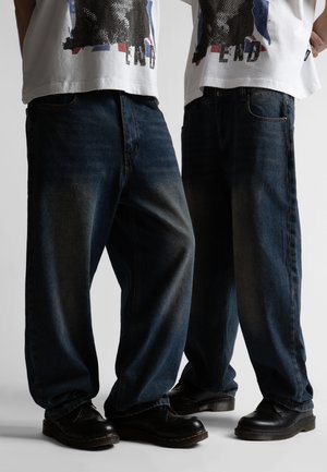 To personer iført løstsiddende mørkeblå jeans, sorte sko og hvide t-shirts med grafik og delvist synligt tryk.