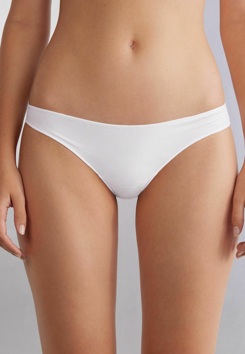 Bílý bavlněný bikini s hladkou texturou, nízkým střihem a minimálním pokrytím. Obsahuje široký, měkký pás pro pohodlí.