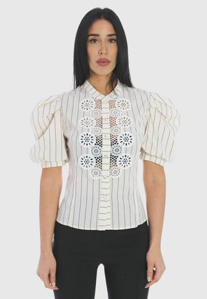 Donna che indossa una camicetta bianca a righe sottili con maniche a sbuffo e una pannellatura frontale con intagli in pizzo floreale, abbinata a pantaloni neri.