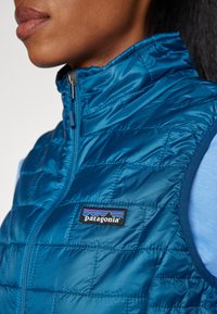 Gilet matelassé bleu en tissu léger et brillant, avec un col haut et un badge noir de Patagonia sur la poitrine gauche.