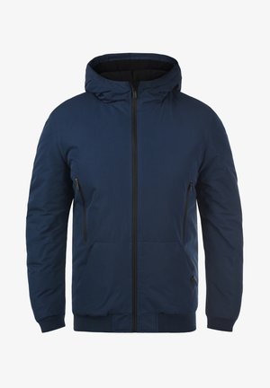 Námořnická modrá zateplená bunda s kapucí, vybavená předním zipem, dvěma zipovými bočními kapsami a ribbenými manžetami na lemu. Měkká textura.