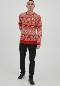 Pull en tricot rouge avec des motifs géométriques blancs, des flocons de neige et des rennes. Col rond avec des poignets et un ourlet côtelés. Associé à un pantalon et des chaussures noires.