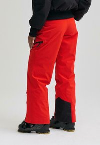 Pantaloni de schi roșii cu un design conic, accente negre la tivuri, buzunare cu fermoar și o textură din țesătură rezistentă la apă.