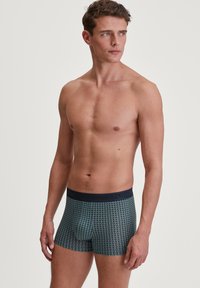 Herenboxershorts in een donkerteal stof met een geometrisch patroon. Elastische tailleband met branding. Nauwsluitend, zachte textuur.
