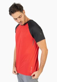 Camiseta atlética roja con mangas raglán negras, tela texturizada, logo en la manga izquierda y un sutil branding en la parte inferior derecha.