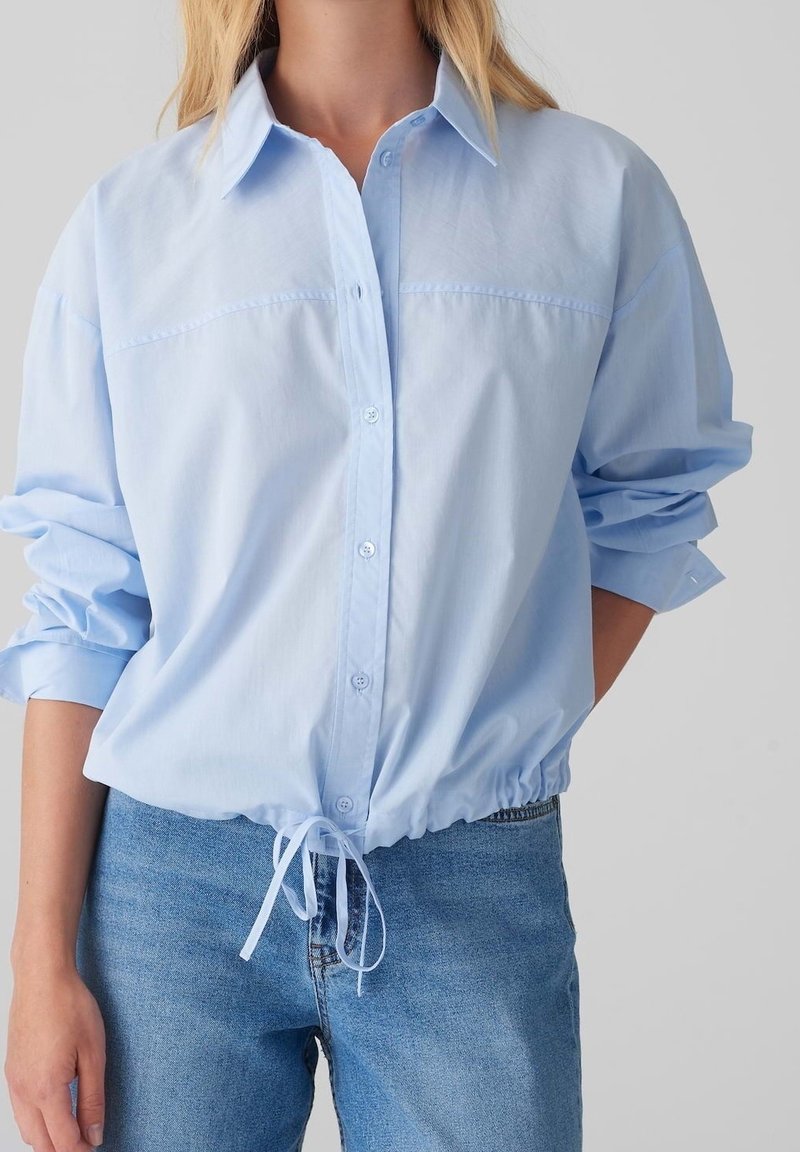Lyseblå skjorte med knapper, der samler sig i taljen, opkrogede ærmer og sømme foran ved brystet. Fremstillet af et glat, let stof.