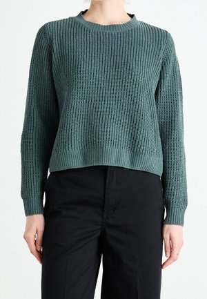 Personne portant un pull en maille bleu sarcelle avec poignets et ourlet côtelés, associé à un pantalon noir taille haute, debout sur un fond blanc.