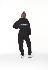 Musta lyhyt huppari, jossa valkoinen "PEGADOR"-teksti, yhdistettynä mustiin samanvärisiin collegehousuihin. Malli pukeutunut valkoisiin urheilukenkiin.