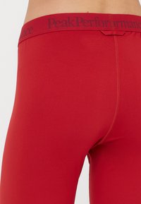 Leggings vermelhos com uma textura suave e cintura elástica, apresentando o logótipo "PeakPerformance" em cinza. Vista em close a focar no design traseiro.