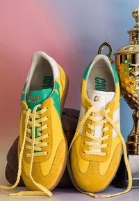 Paire de chaussures de sport jaunes et vertes avec des lacets défaits, placées devant un ballon de football marron et un trophée en or sur un fond rose-bleu.