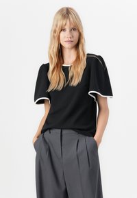 Femme blonde aux cheveux ondulés portant un haut noir à manches courtes avec des bordures blanches et un pantalon plissé gris taille haute ample, les mains dans les poches.