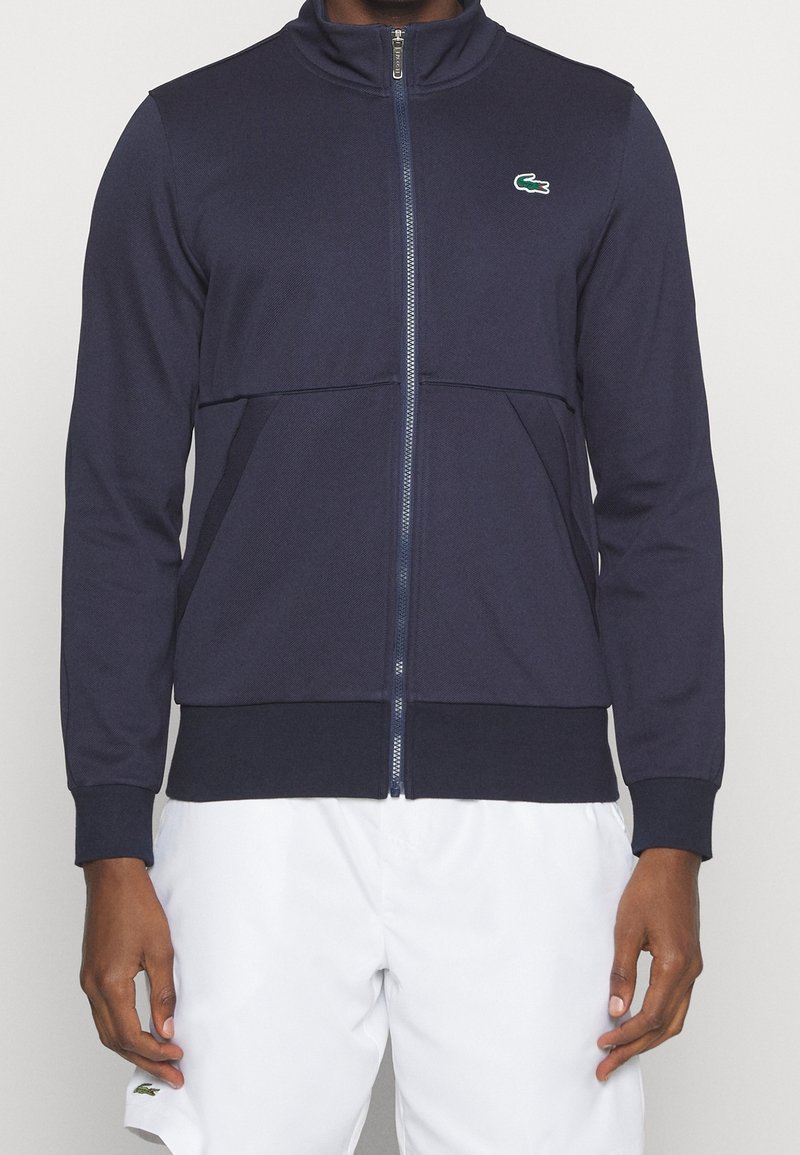 Dunkelblaue Zip-Jacke aus strukturiertem Stoff, mit hohem Kragen und grünem Markenlogo auf der linken Brust. Mit Seitentaschen.