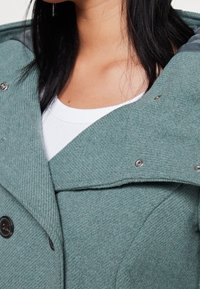 Groene wollen mix jas met een gestructureerde afwerking, grote kraag, drukknoopsluiting en gelaagd ontwerp. Eronder is een witte geribbelde top zichtbaar.