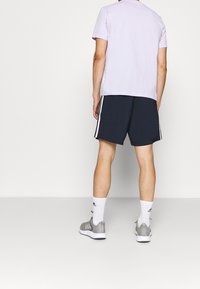 Camisa corta de manga corta color púrpura claro combinada con pantalones cortos deportivos azul marino con franjas laterales blancas. Usada con zapatillas deportivas grises y calcetines blancos.