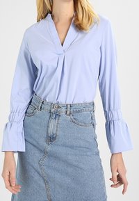 Blusa azzurra chiara con scollo a V e parte frontale plissettata, caratterizzata da maniche lunghe con polsini arricciati. Abbinate a una gonna di jeans, che presenta uno spacco frontale.
