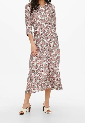 Mujer con un vestido cruzado de paisley en blanco, rosa y marrón, de manga tres cuartos y sandalias de tacón, de pie sobre un fondo liso.