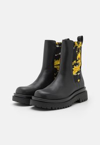 Botas de tobillo de cuero negro con suelas gruesas y paneles laterales elásticos que presentan un patrón floral barroco amarillo sobre un fondo negro.