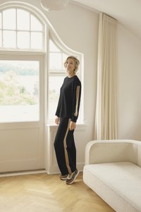 Schwarzes Loungewear-Set mit langen Ärmeln und Hose mit goldenen Seitenstreifen. Der Stoff wirkt weich; das Outfit wird mit lässigen Sneakers kombiniert.
