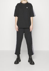 Černé oversized tričko s bílými pruhy, krátkými rukávy a logem. Černé tepláky s bočními pruhy a masivními černými botami.