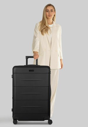 Zwarte harde koffer met gestructureerd oppervlak, horizontale groeven, uitschuifbare handgreep en vier wielen, geplaatst naast een persoon in lichte kleding.