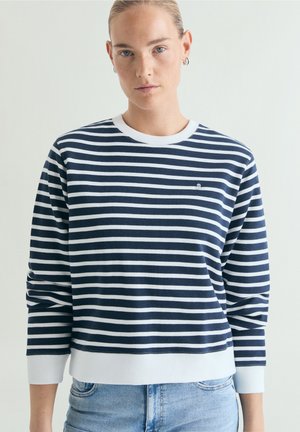 Donna con capelli biondi che indossa una maglia a maniche lunghe a righe blu navy e bianco e jeans azzurri, in piedi contro uno sfondo semplice.