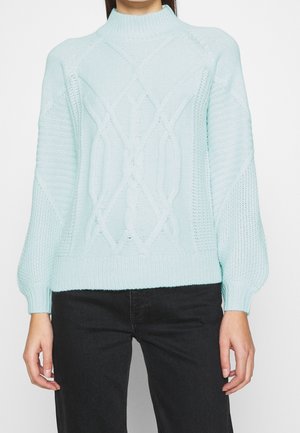 Nainen, joka pukeutunut vaaleansiniseen palmikkoneulepaitaan, jossa on ribbireunukset hihansuissa, ja mustiin housuihin yksinkertaista taustaa vasten.