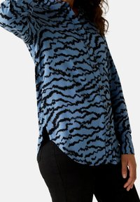 Camicia button-up in tessuto blu con un motivo a onde nere. Maniche lunghe con spacchi laterali; texture liscia e vestibilità strutturata.