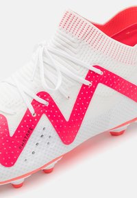 Scarpa da calcio bianca con design zigzag rosa, lacci bianchi e fodera interna rosa, che mostra un materiale a maglia testurizzata e piccoli fori di ventilazione.