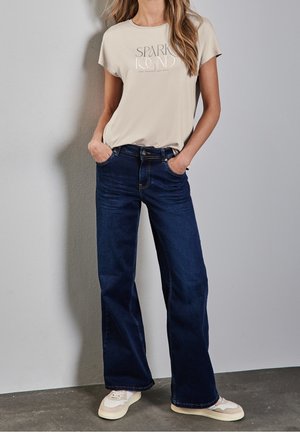 Vrouw draagt beige "Spark Found" t-shirt, donkerblauwe wijd uitlopende jeans en beige sneakers, staand met handen in zakken tegen een grijze muur.